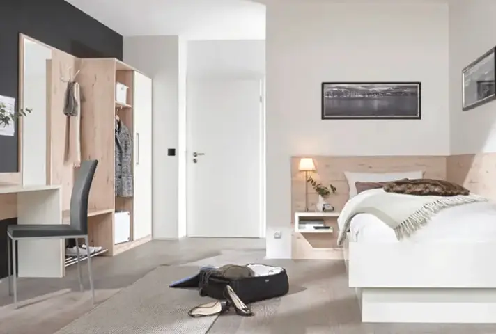 Schlafzimmer mit Garderobenschrank und offenem Fach links, weissem Bett mit Nachttischnische rechts, dazwischen weisse Tür, vorn offener Koffer und Schuhe auf Teppich.